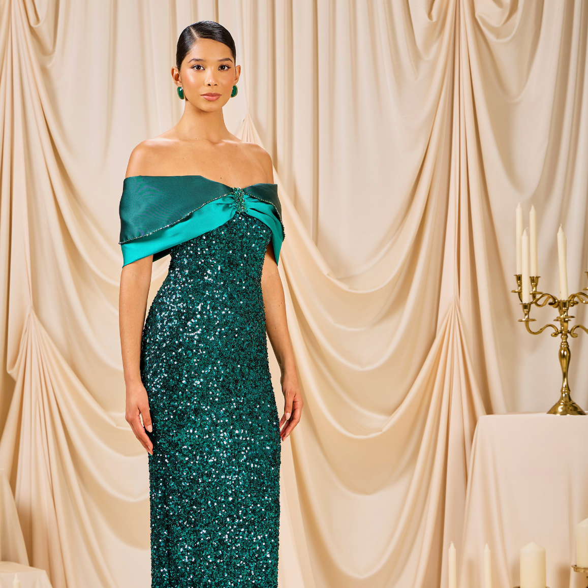 Woman in a green sequin dress standing in front of beige curtains with a candelabra in the background.